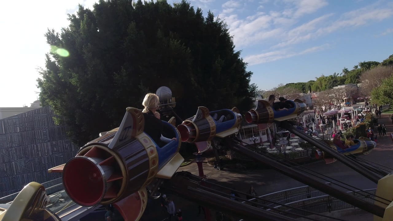 Several rocket-shaped cars appear at different heights over Disneyland California, with riders in the cars and guests moving through the walkways below.