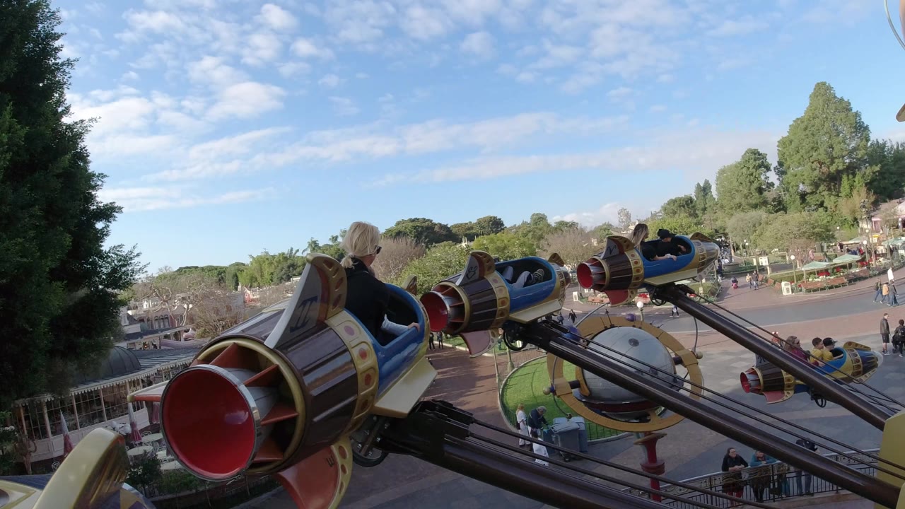 Rocket cars arc over trees, walkways, and a circular ride structure.