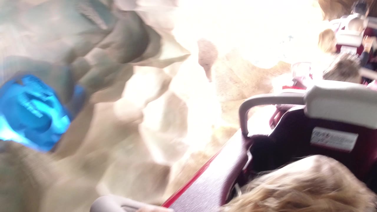 At Disneyland California, the camera passes close to a bright white tunnel wall with a blue circular light feature while riders and coaster restraints fill the right side of the frame.