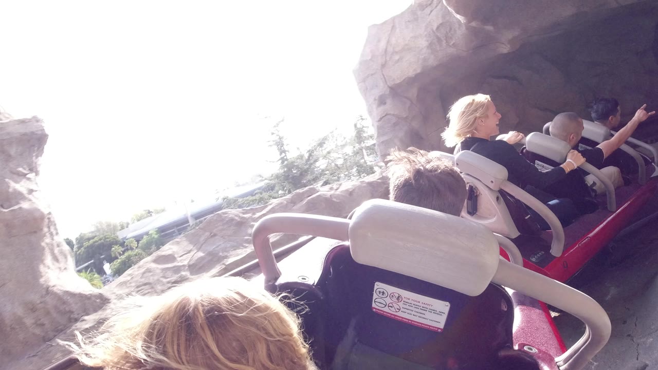 Disneyland California rollercoaster riders seen from behind as a red car enters a rock-lined tunnel with bright daylight at the opening.