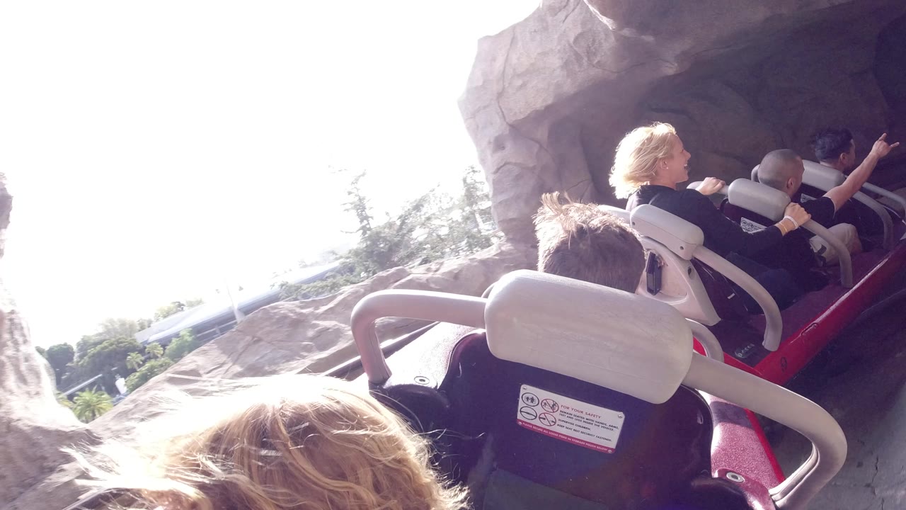 Riders in a red coaster car pass through a rock tunnel at Disneyland California, with a bright exterior opening and seat restraints close to camera.