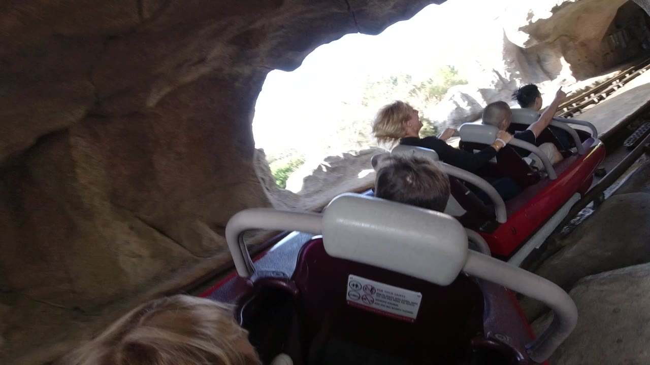 A red rollercoaster car with several riders moves through a darker rock tunnel, with rails visible on the right and bright daylight behind the cave edge.