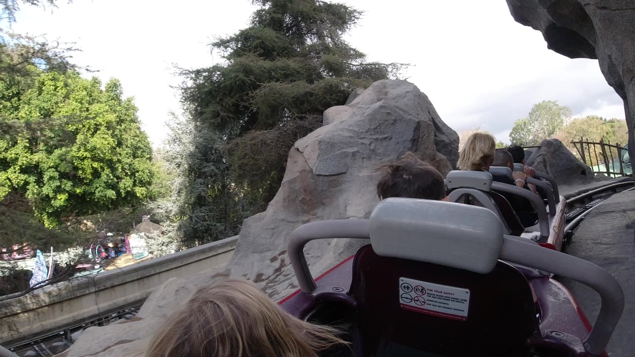 The Disneyland California ride POV continues behind seated guests, with gray seat backs, a curving track, artificial rocks, and green trees in view.