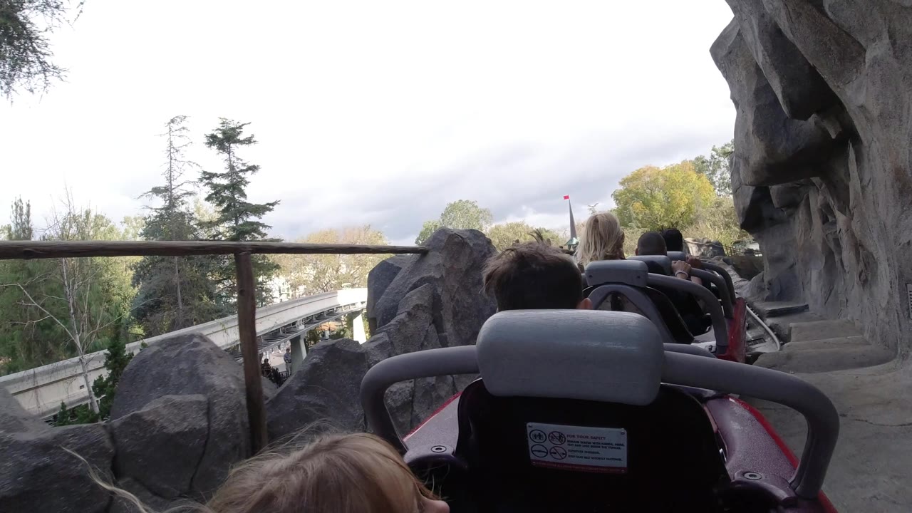 A Disneyland California coaster view shows passengers ahead, gray rock walls at both sides, a metal rail crossing the frame, and park trees beyond.