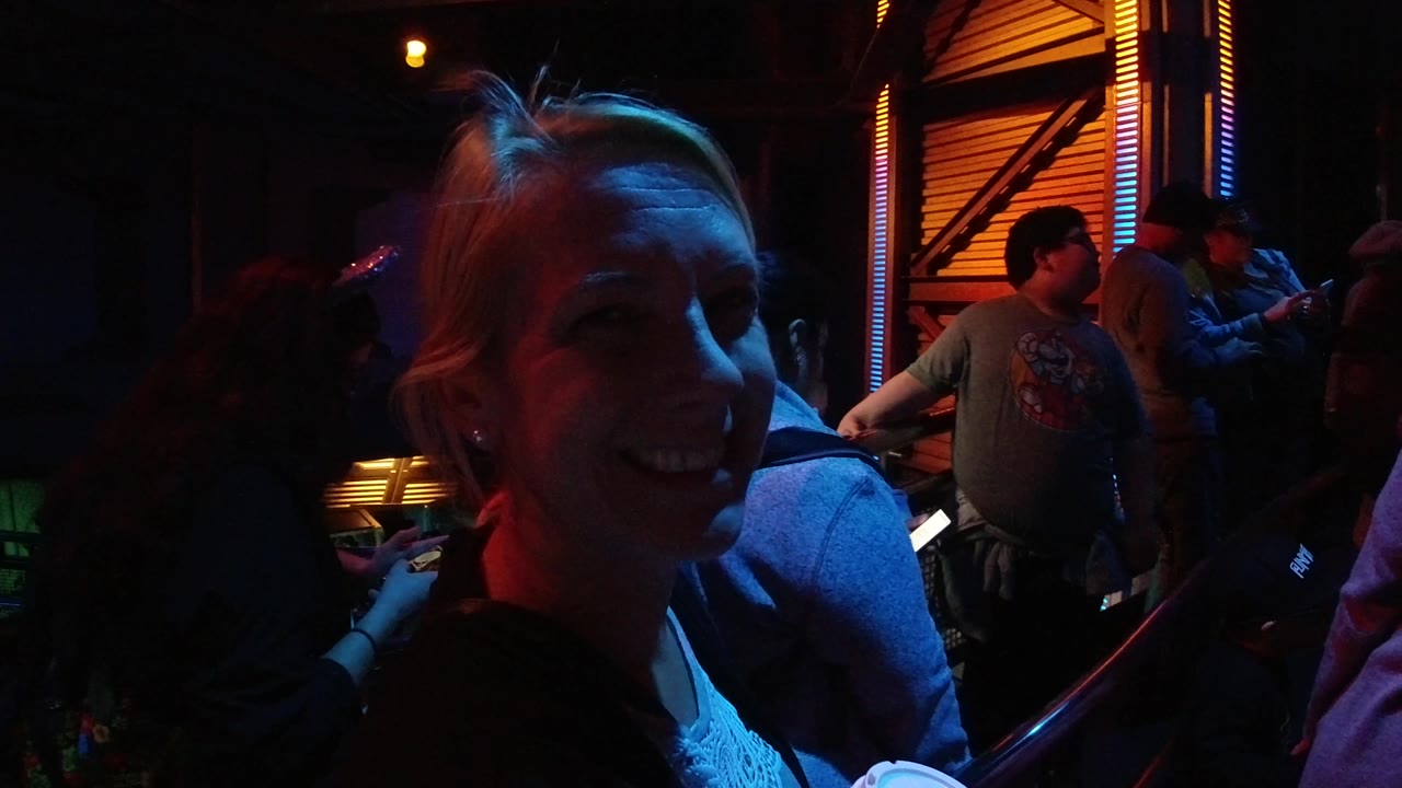 At Disneyland California, the close foreground woman smiles toward the camera while a man in a graphic shirt and other visitors are visible under orange and blue lighting.
