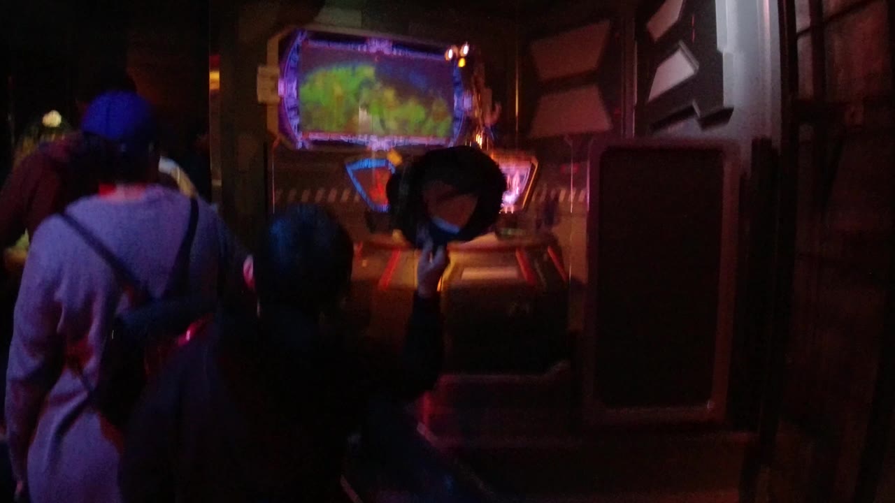 At Disneyland California, guests stand in a dark indoor queue facing a glowing display, with a man and child seen from behind under red and blue attraction lighting.