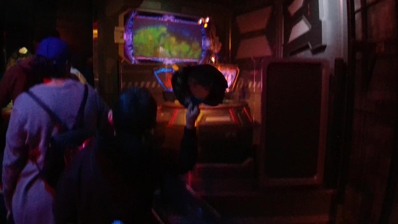 People wait in a dark indoor queue with a bright screen or display in front of them.
