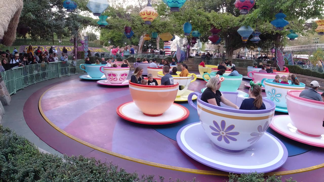 Wide view of the Disneyland California teacup ride with several pastel cups carrying adults and children, queue railings at left, and hanging lanterns above the platform.