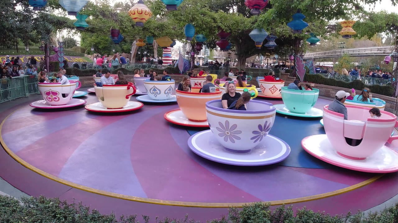 Wide Disneyland California amusement ride view with a purple cup near the foreground, mint and pink cups to the right, seated riders, spectators behind railings, trees, and colorful lanterns.