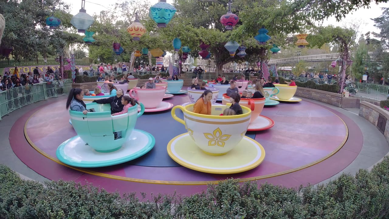 A wide view of the Disneyland California cup-and-saucer ride shows riders in turquoise, yellow, and pink cars, with a queue area and trees behind them.