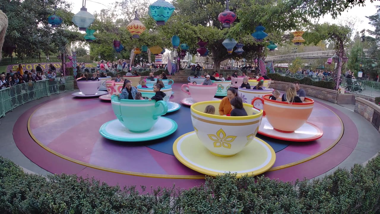 The Disneyland California ride is seen with a turquoise cup near center, yellow and orange cups to the right, hanging lanterns overhead, and guests seated inside the cars.