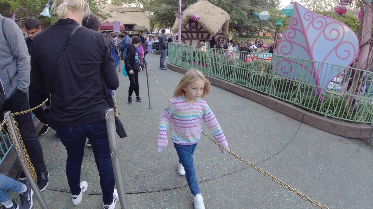 At Disneyland California, a little girl in a striped sweater walks beside a chain barrier while adults and queued visitors fill the walkway and railings behind her.