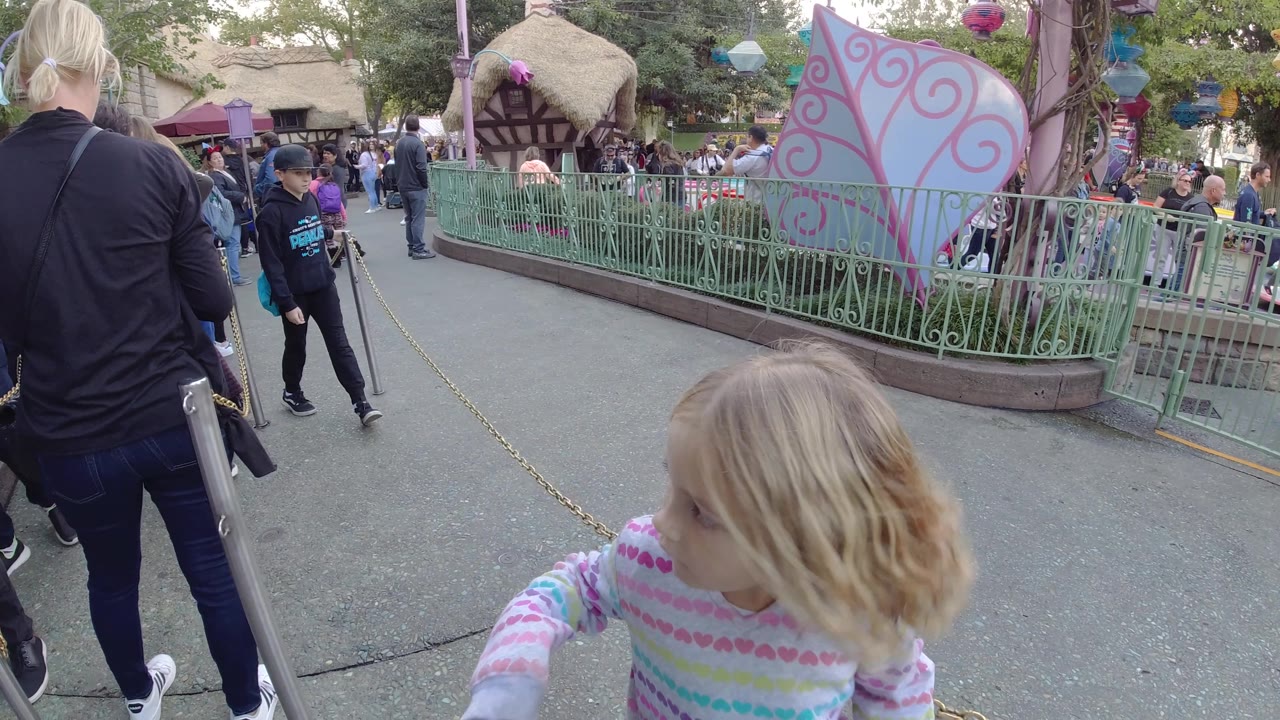 At Disneyland California, the girl passes close to the right foreground while a boy in a dark cap and hoodie approaches from the midground near the chain barrier and railings.
