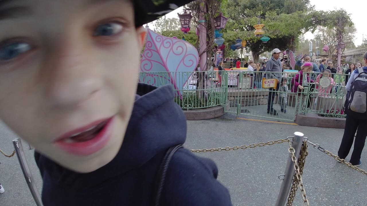 At Disneyland California, a boy's face fills the left foreground while green railings, pastel ride panels, chain barriers, trees, and visitors are visible behind him.