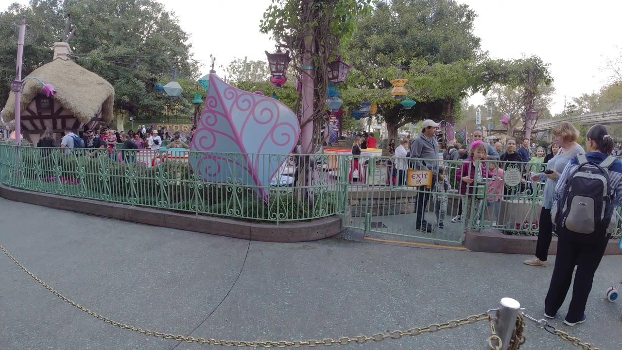 At Disneyland California, the frame widens to show green queue railings, pastel ride panels, a thatched-looking themed structure, trees, and many park visitors along the walkway.