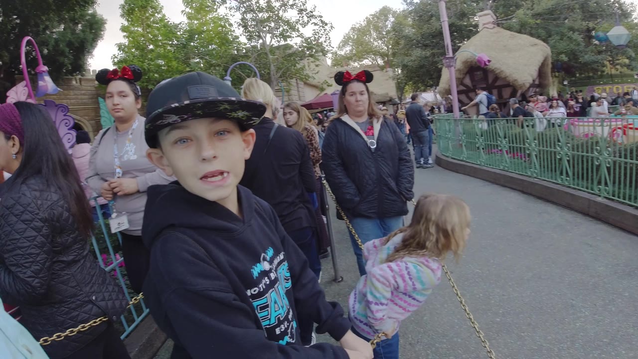 A boy in a black cap looks toward camera at Disneyland California as a little girl in a striped top, adults, queue chains, green railings, and pastel ride-area details surround him.