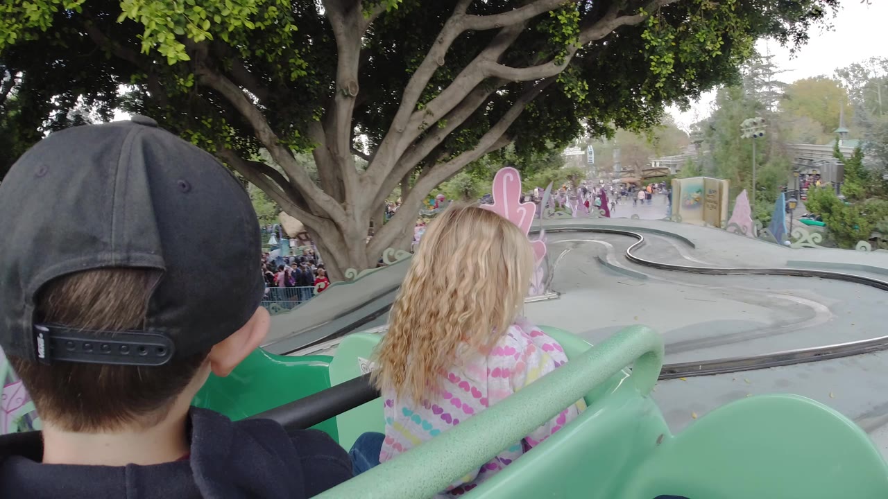 At Disneyland California, children sit in a green roller coaster car with a boy in a black cap in the foreground and a blonde child ahead facing the track.