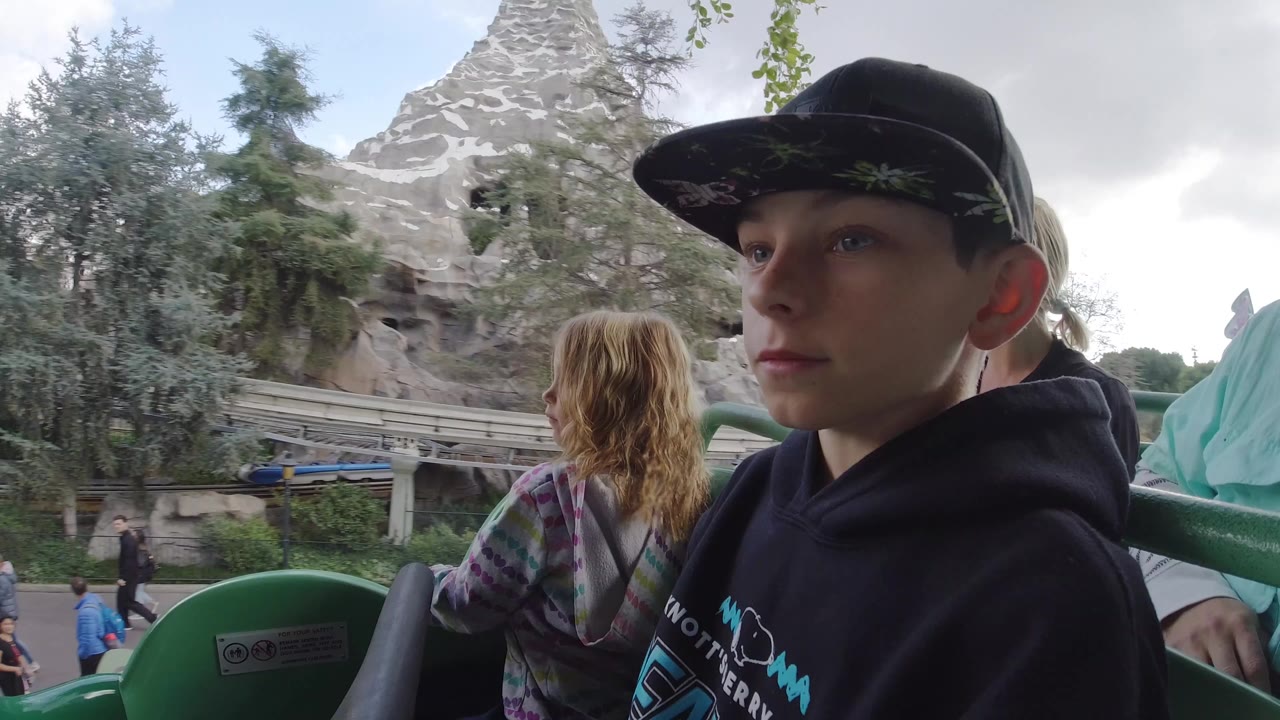 At Disneyland California, a boy in a black cap and hoodie sits in the foreground of a green roller coaster car while a girl faces forward and a rocky mountain backdrop fills the rear of the frame.