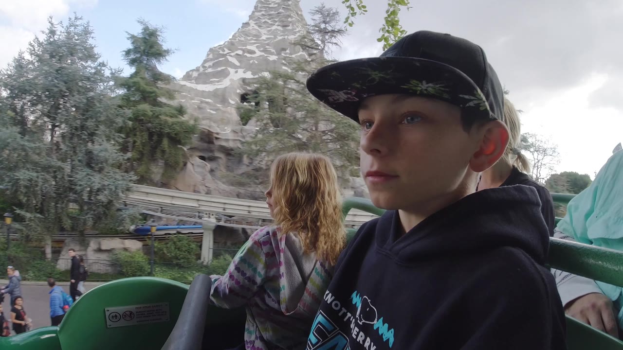 At Disneyland California, the boy remains close to camera in the coaster car while the girl looks ahead and the rocky ride structure, track, trees, and scattered visitors sit behind them.