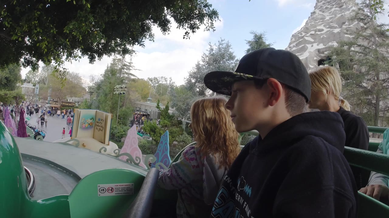 At Disneyland California, the view shifts to a side angle of the boy and girl in the green coaster car, with a busy walkway, pastel decorative set pieces, trees, and park structures visible below.