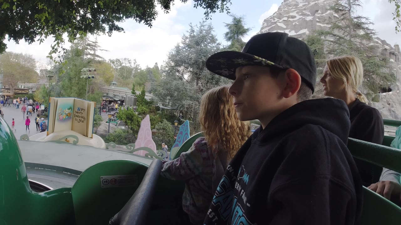 Boy in profile rides beside a girl and adult woman with a crowd and decorative theme park scenery below.