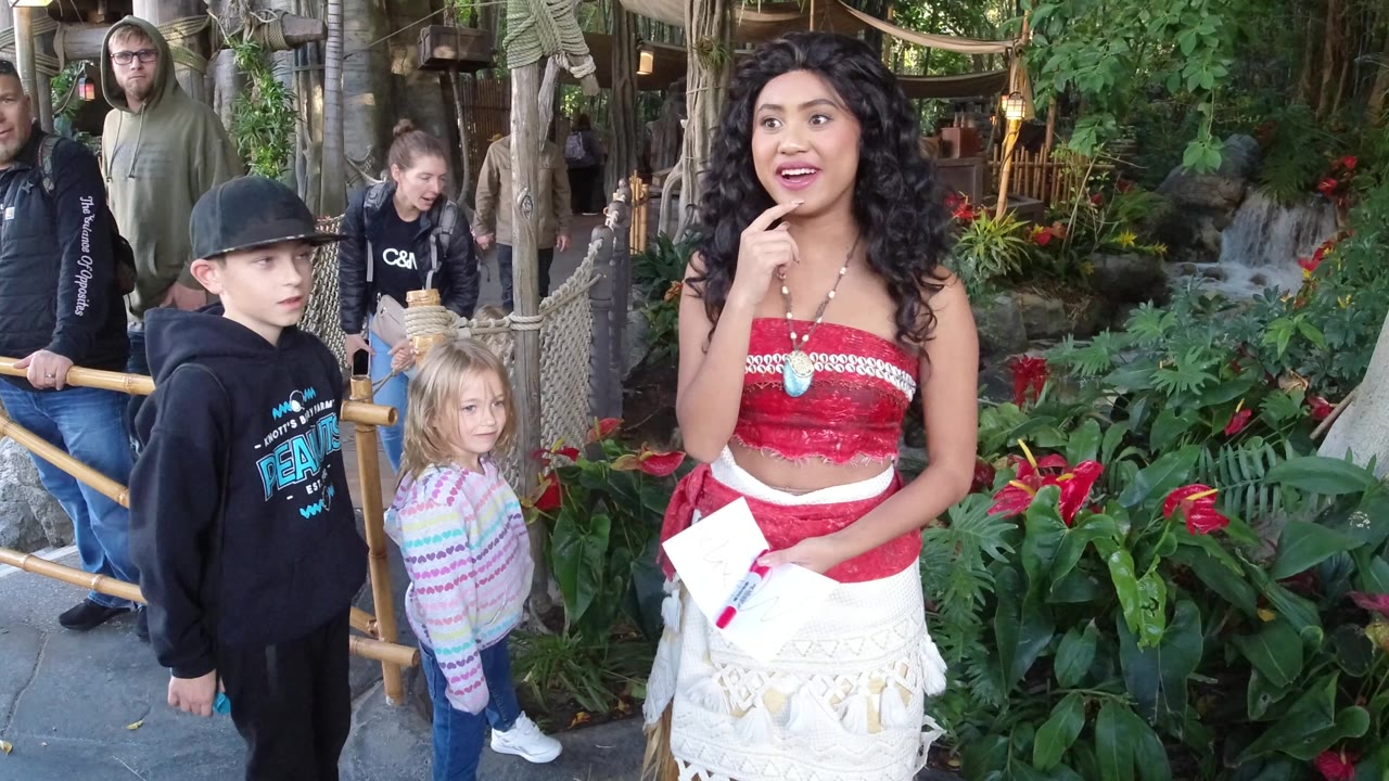 At Disneyland California, a boy in a black hoodie and a young girl stand near a costumed woman holding papers beside bamboo railings and dense planting.