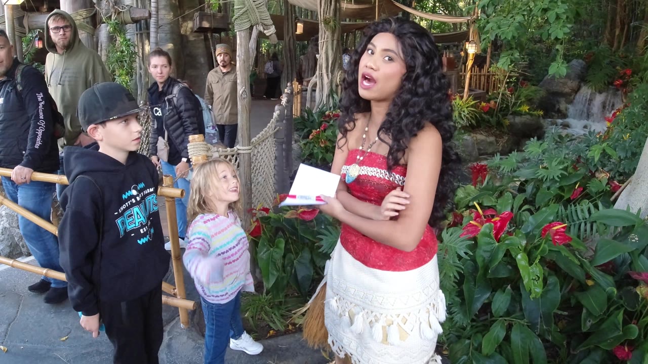 At Disneyland California, the costumed woman holds papers and turns toward the children, with the young girl looking up near the bamboo rail and planted backdrop.