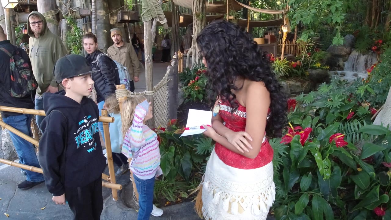 Children face a costumed woman from behind beside a planted walkway.