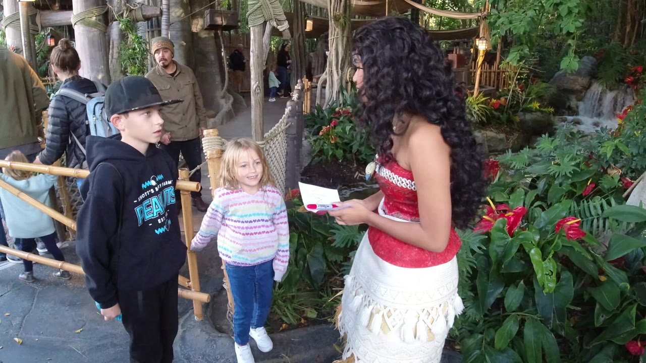 At Disneyland California, a woman dressed as Moana stands beside tropical planting and speaks with a boy in a black cap and a blonde girl near a bamboo railing.