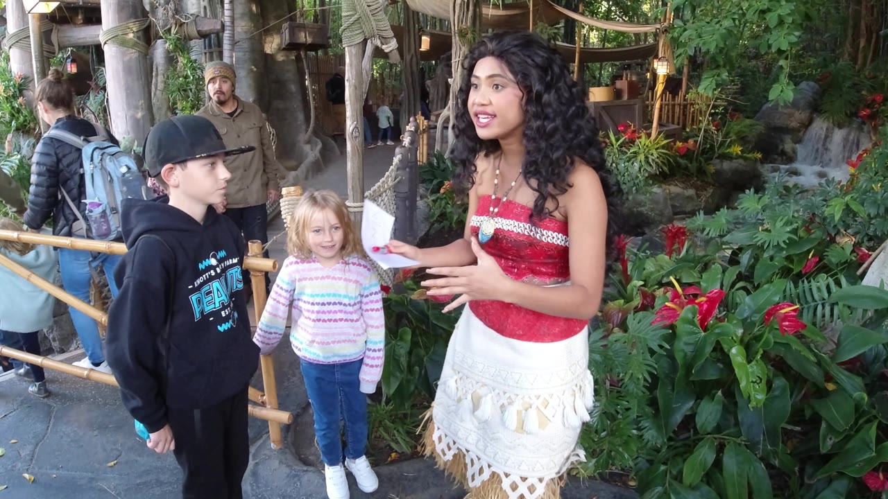 At Disneyland California, the Moana-costumed woman gestures with both hands while two children stand near the bamboo rail and themed greenery.