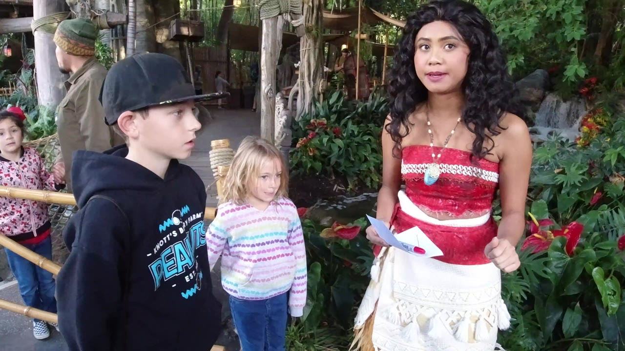 At Disneyland California, a boy in a black cap and hoodie and two girls stand near a costumed performer in red and white beside tropical plants and a water feature.