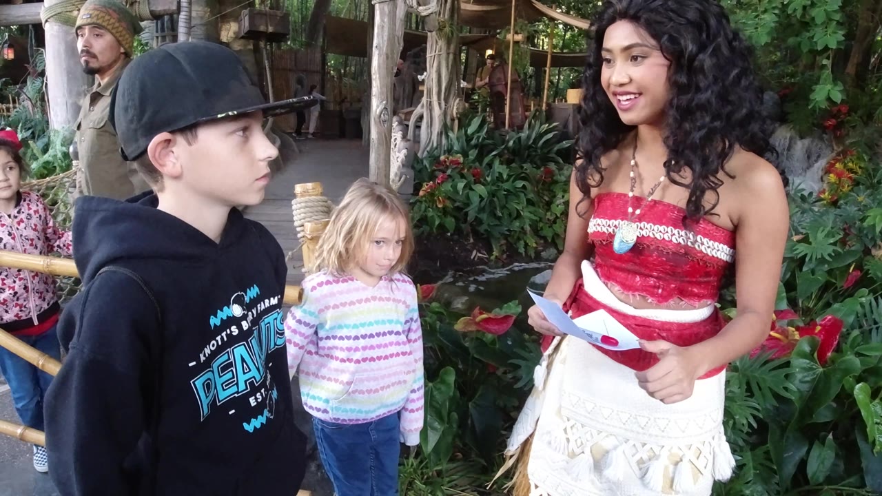 At Disneyland California, the performer smiles toward the children while the boy looks to the side and the blond girl stands between them near dense tropical planting.