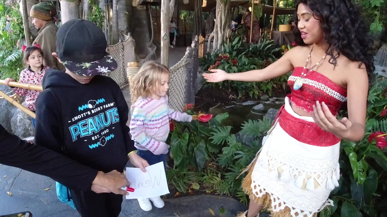 At Disneyland California, the boy looks down at a white paper near the costumed woman as the young girl stands beside tropical plants and bamboo railings.