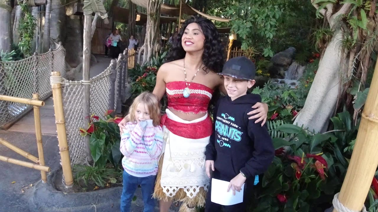 At Disneyland California, a costumed woman stands with two children beside bamboo railings, rope netting, green plants, and red flowers at the start of the clip.
