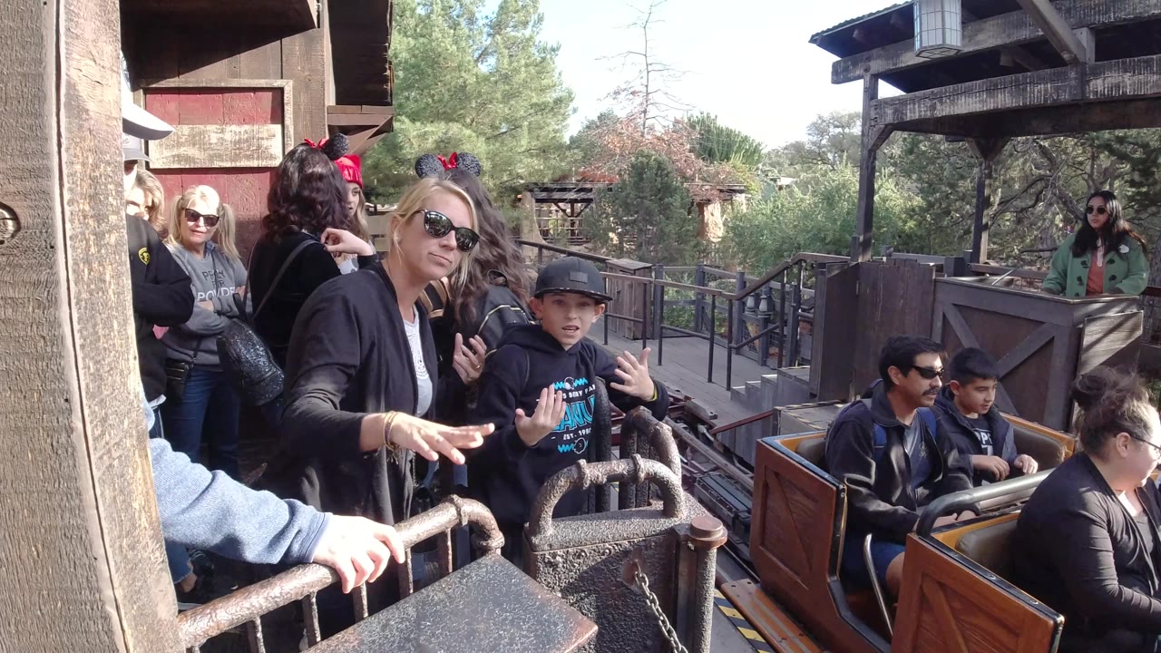 At Disneyland California, a woman in sunglasses and a child in a black cap stand near the station rail while coaster cars with riders move farther along the track.