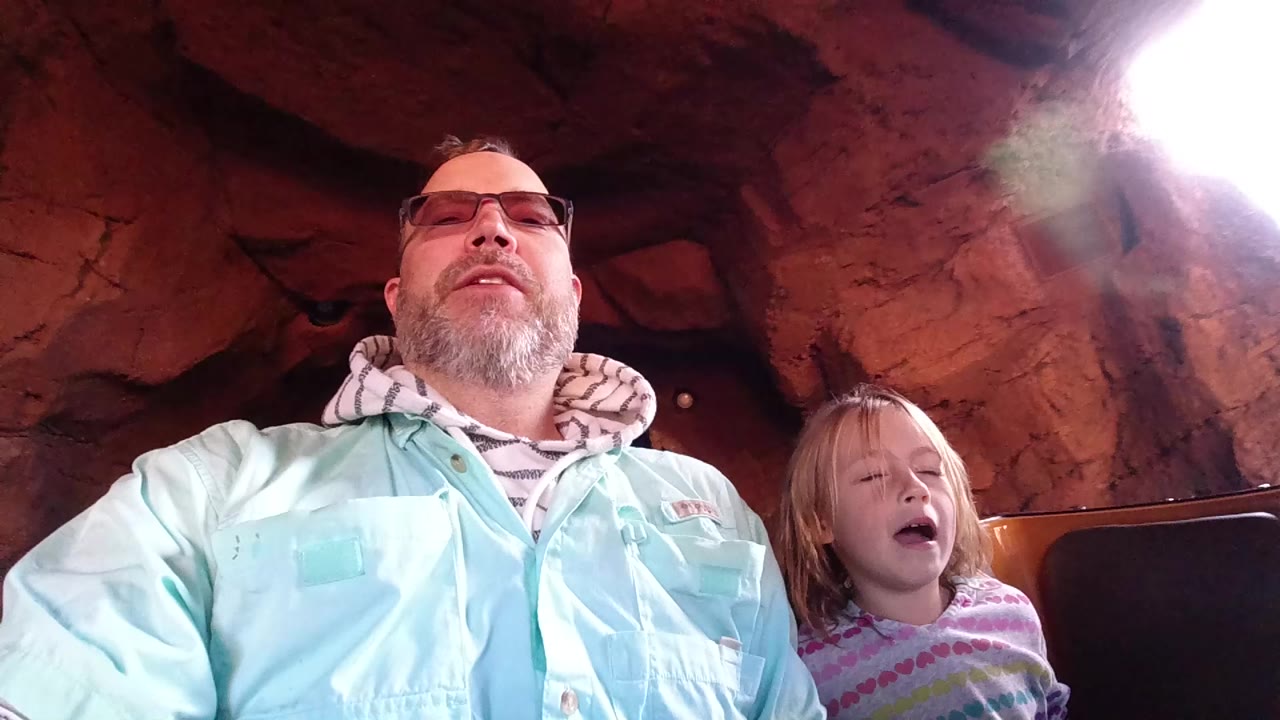 At Disneyland California, a father in glasses and a young daughter sit side by side in a log flume boat beneath reddish cave rock, with bright light flaring at the tunnel opening.