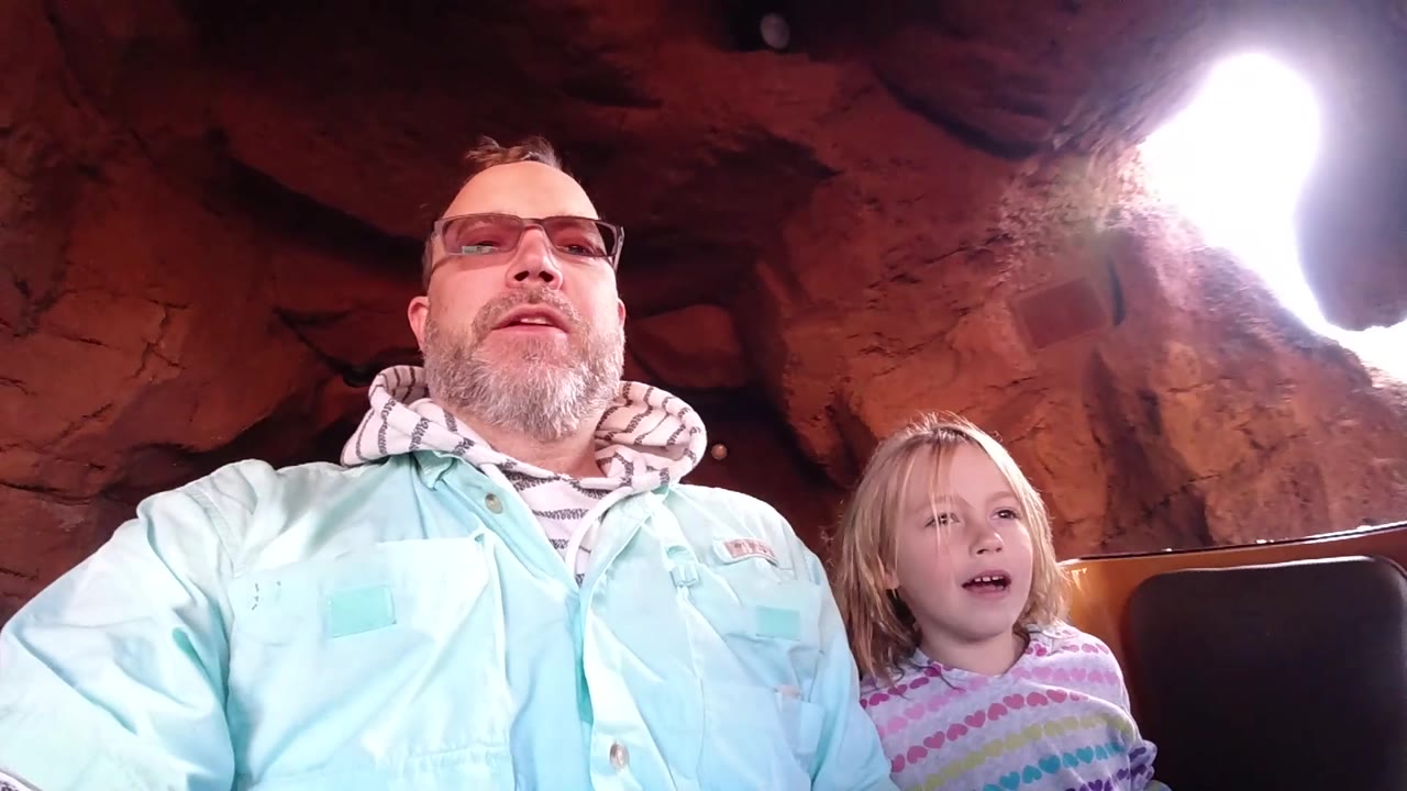 At Disneyland California, the father and daughter are seated under dark reddish cave rock as a white blown-out tunnel opening enters from the upper right of the frame.