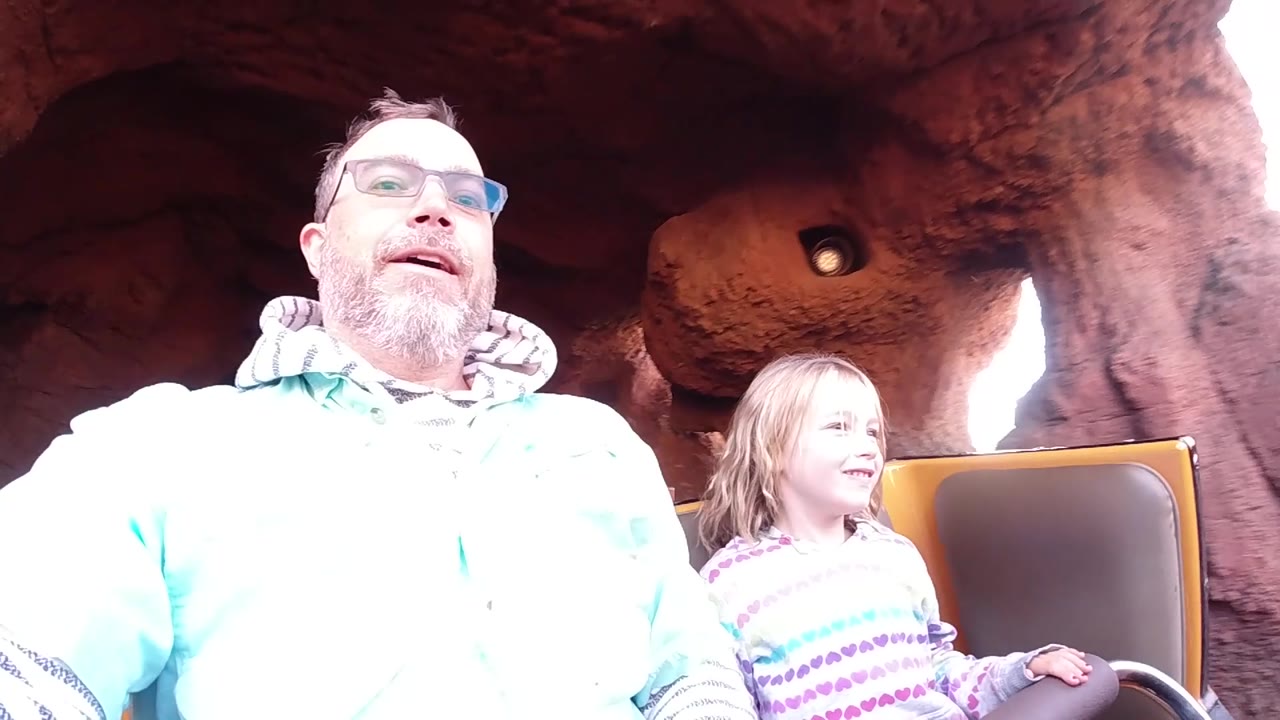 At Disneyland California, the child smiles beside the man in the coaster car, with reddish artificial rock, a small overhead fixture, and washed-out daylight visible behind the seat.