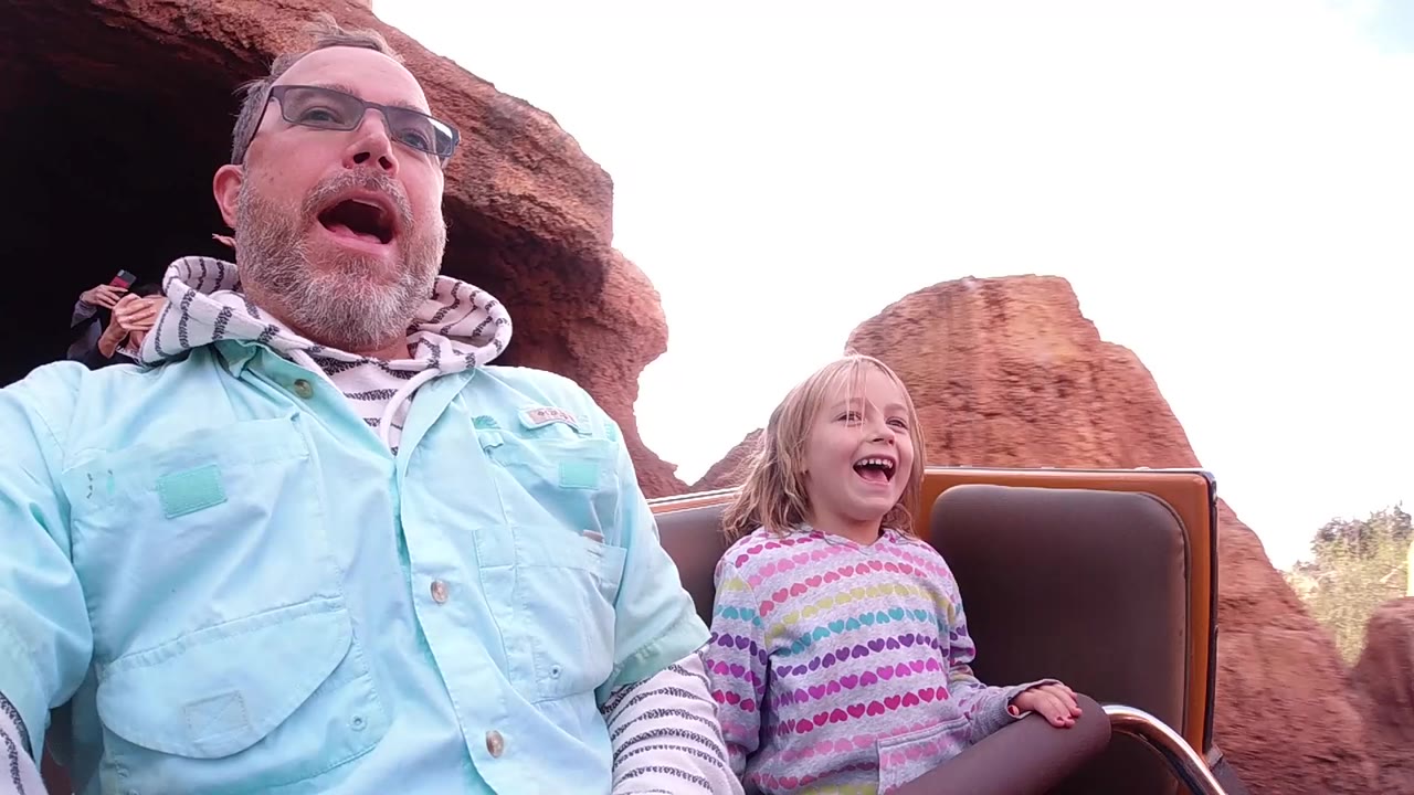 At Disneyland California, a father in glasses and a young daughter laugh in a brown roller coaster car with red rock scenery behind them at the start of the clip.