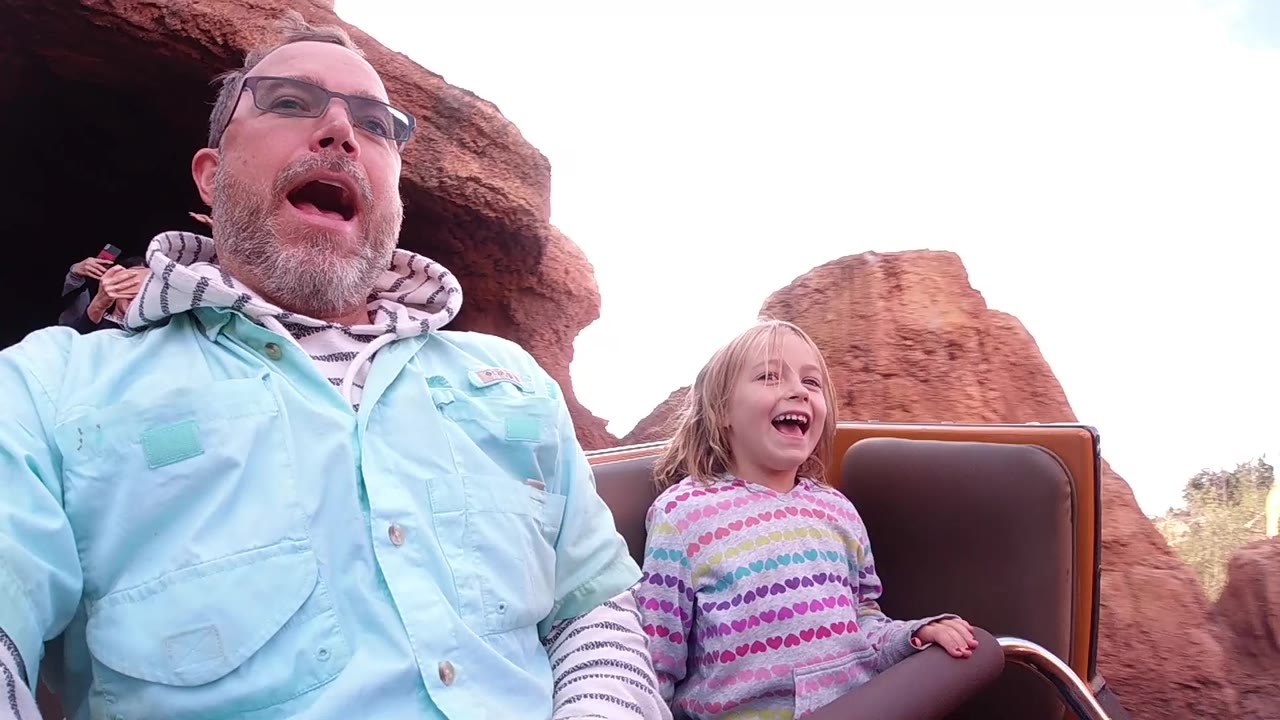 Father and daughter smiling on a roller coaster with bright sky and red rocks behind them.