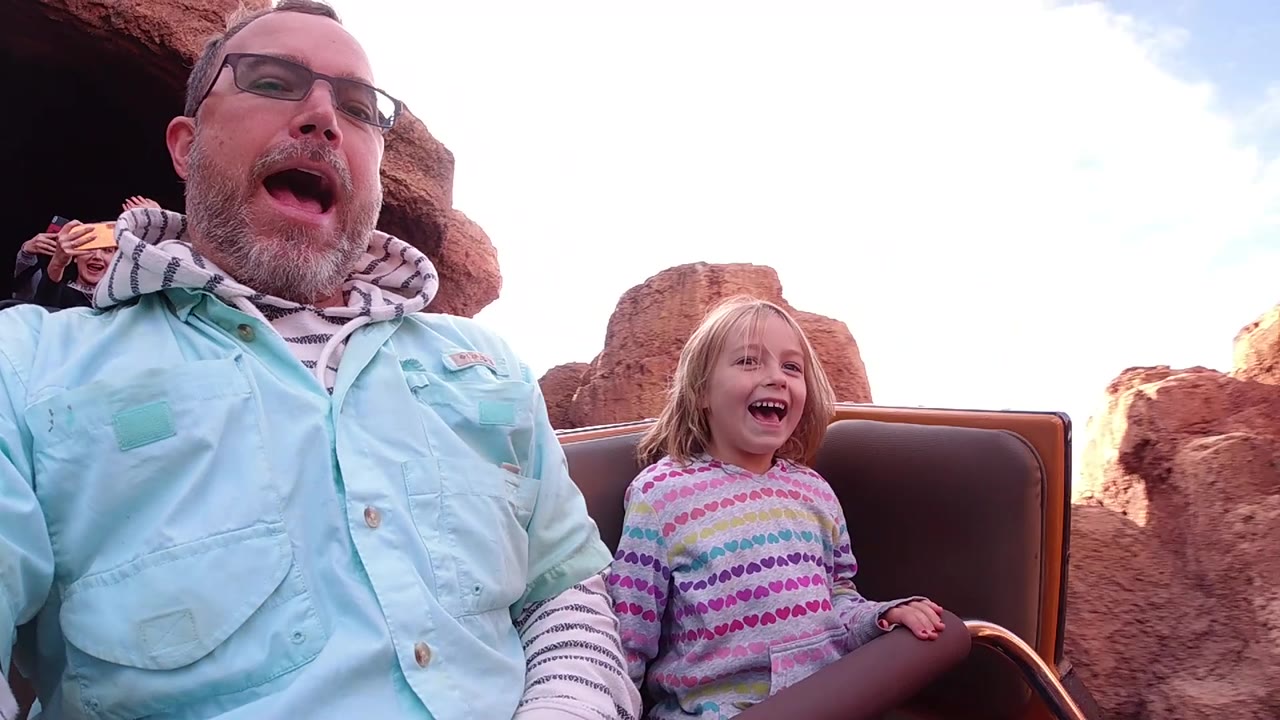 At Disneyland California, the riders lean through a tight wide-angle frame as the daughter laughs near the seat edge and reddish rock formations close in behind the car.