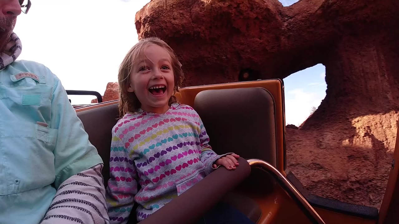 A young girl laughs in a brown ride seat beside an adult at Disneyland California, with red rock scenery behind her.