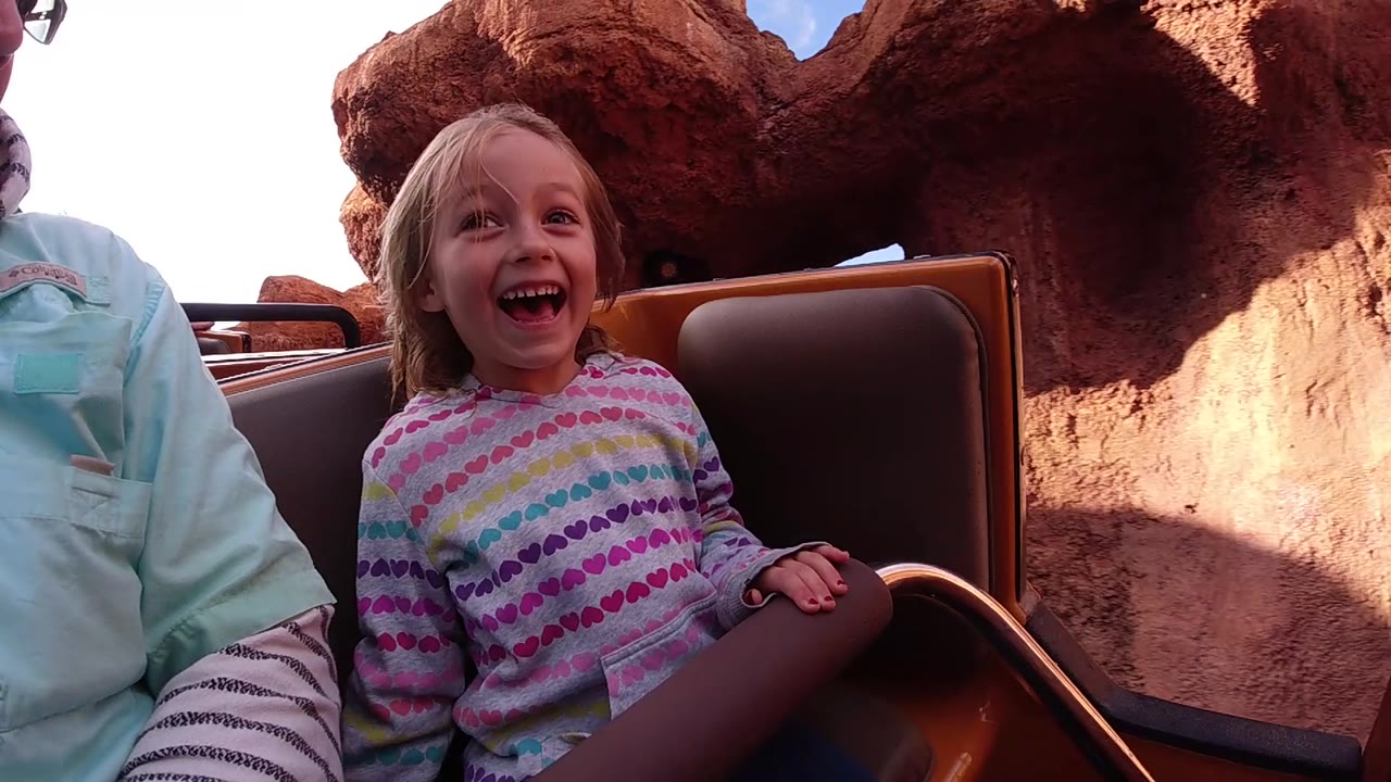 A laughing child sits beside an adult in a ride vehicle at Disneyland California, framed against close red rock walls and bright sky.
