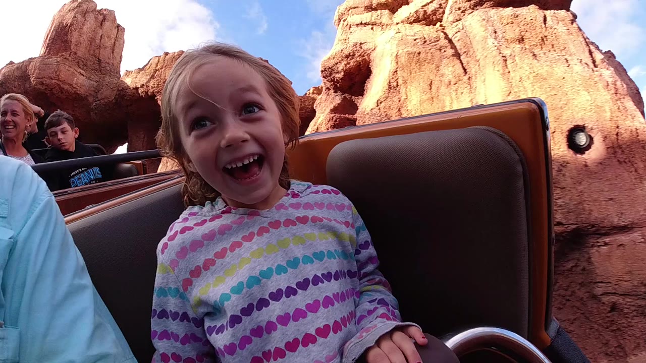 A young child smiles widely in the foreground of a roller coaster car at Disneyland California, with riders, brown seat backs, red rock scenery, and blue sky behind her.