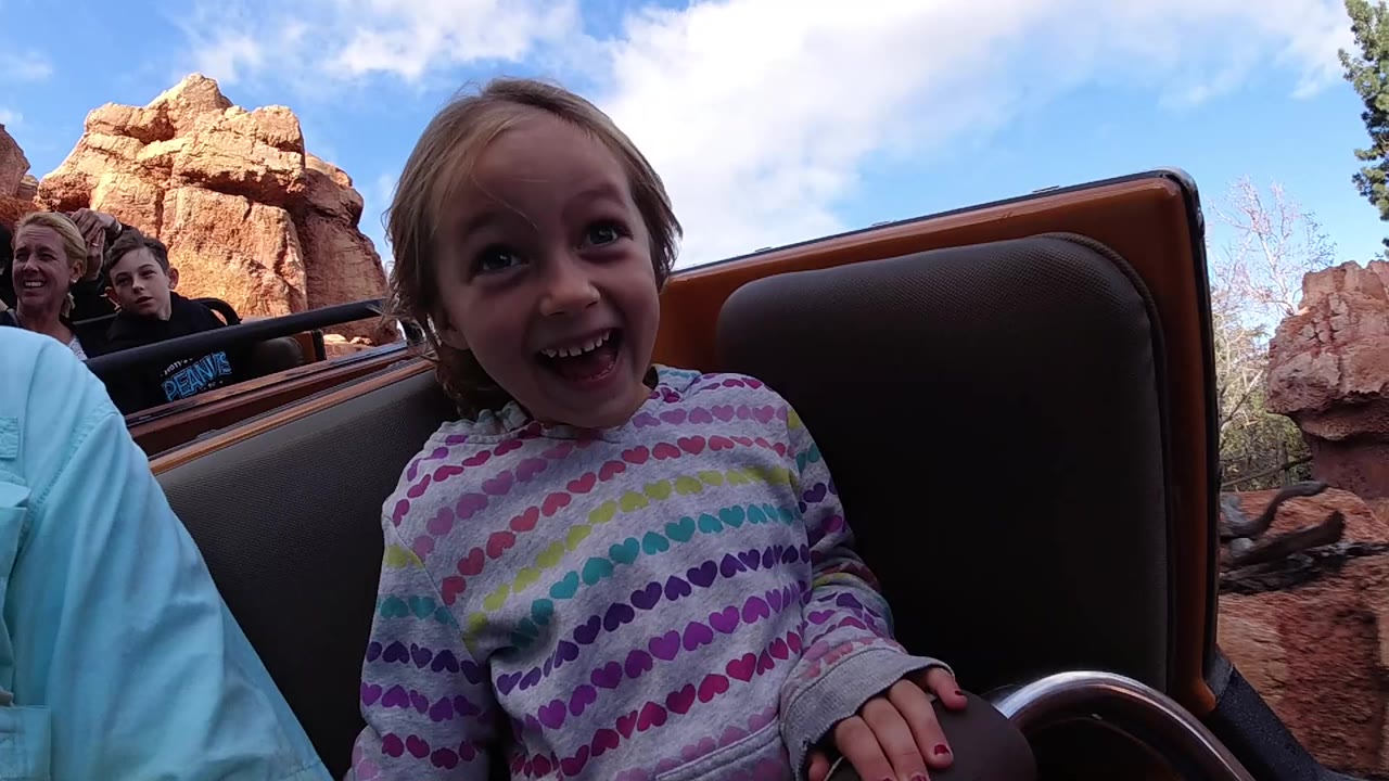The child remains centered in the Disneyland California ride car, grinning beside a tall brown seat back while riders and red rock formations fill the background.