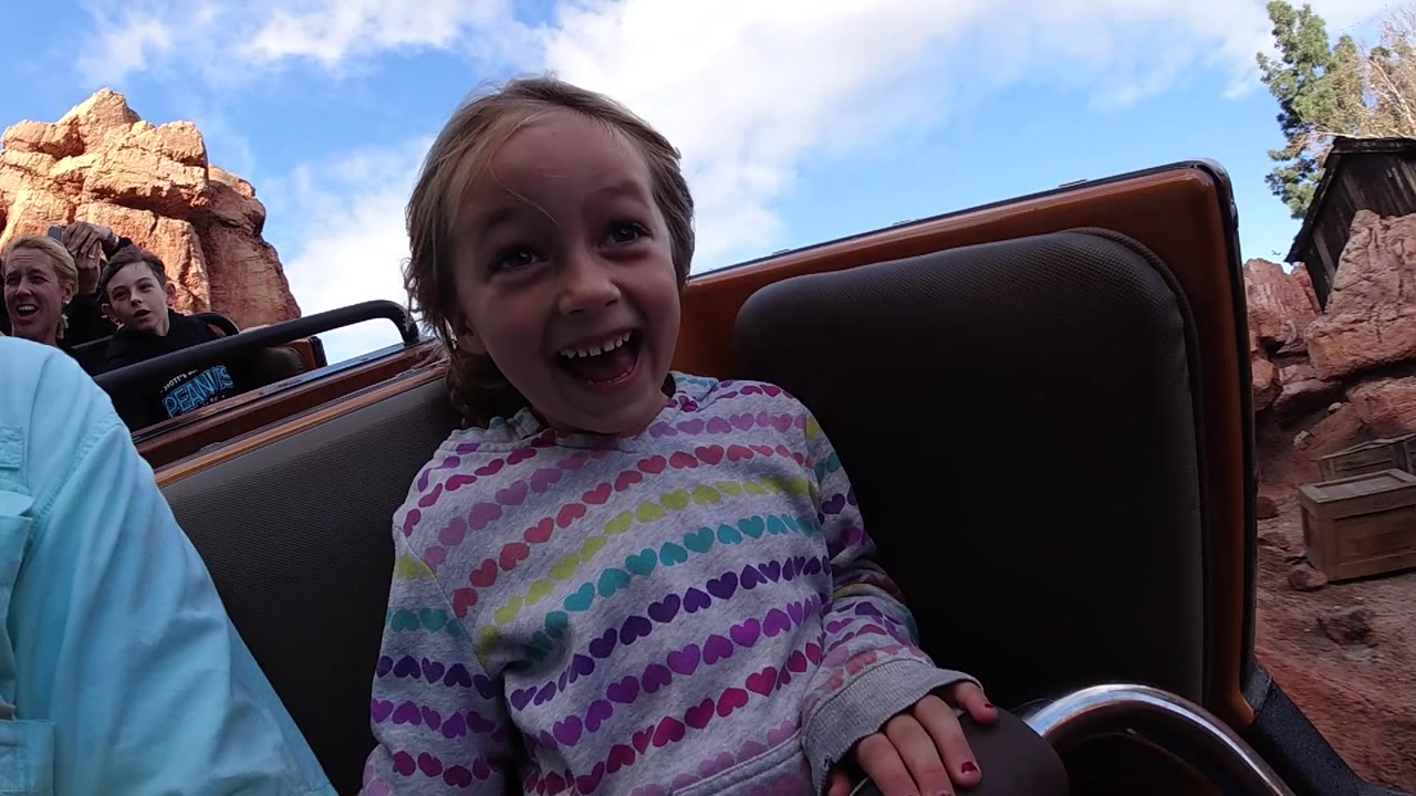 A happy child in a patterned shirt sits low in the Disneyland California ride car as reddish rock scenery, a dark structure edge, and riders behind remain visible.
