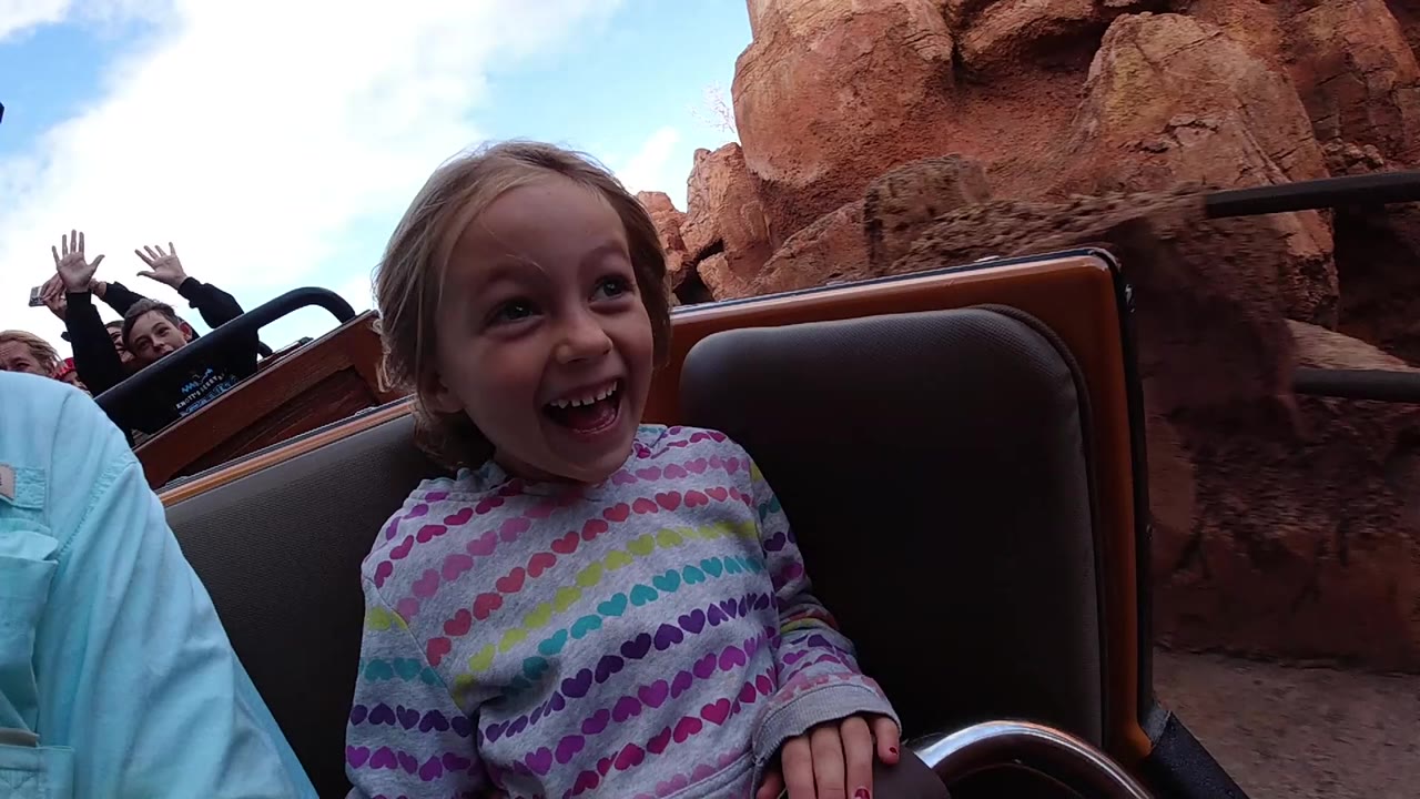 At Disneyland California, a young white girl smiles in the foreground of a roller coaster car while other riders and red rock ride scenery appear behind her.
