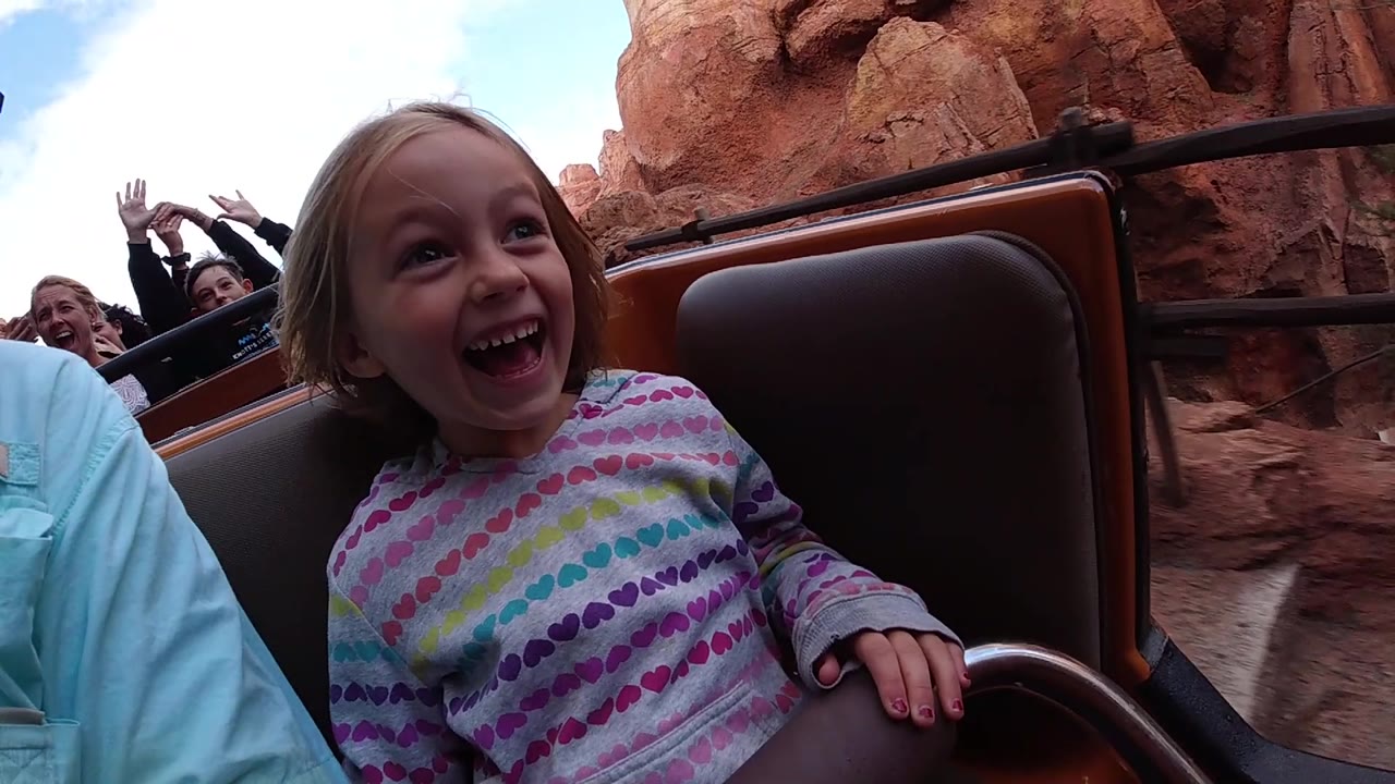 Girl grinning in foreground with raised hands from riders in the background.