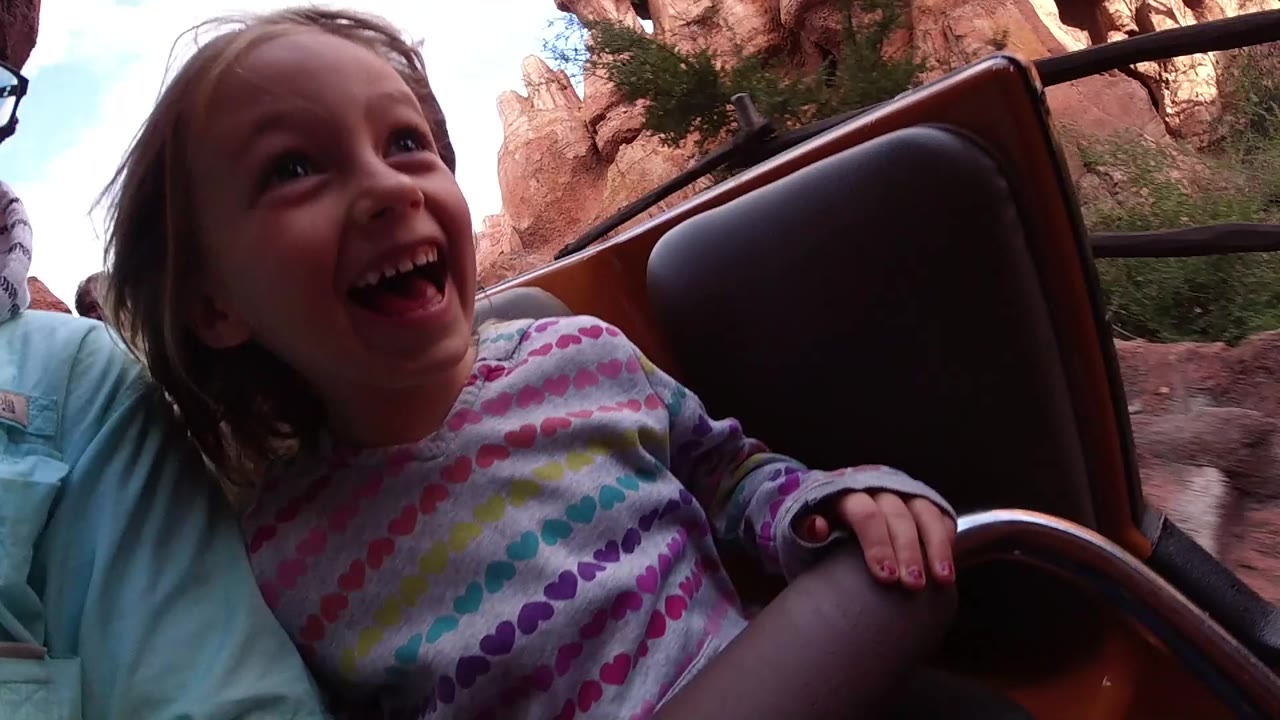 At Disneyland California, a young girl smiles in the foreground of a ride vehicle beside an adult, with red rock walls and a brown seat back visible behind her.