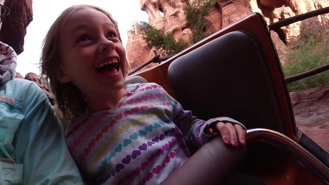 At Disneyland California, the girl faces upward laughing, with more of the rocky canyon backdrop and greenery visible behind the ride seat.